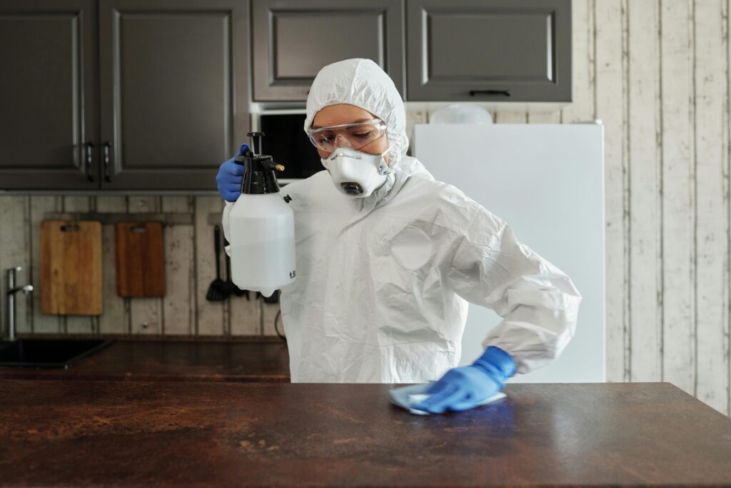A person in protective clothing disinfects a kitchen counter indoors, focusing on safety and cleanliness.