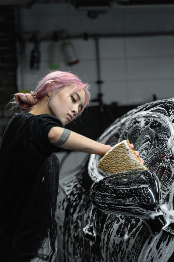 A young woman with dyed hair and tattoos cleaning a car with soap and sponge in a garage.